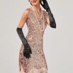 Photo d'une femme portant une robe rose entièrement pailletée style années 20 avec franges roses en bas, un collier de perles blanches, de longs gants noirs satinés, un serre-tête avec plume noire et boucles d'oreilles noires, le tout sur un fond gris