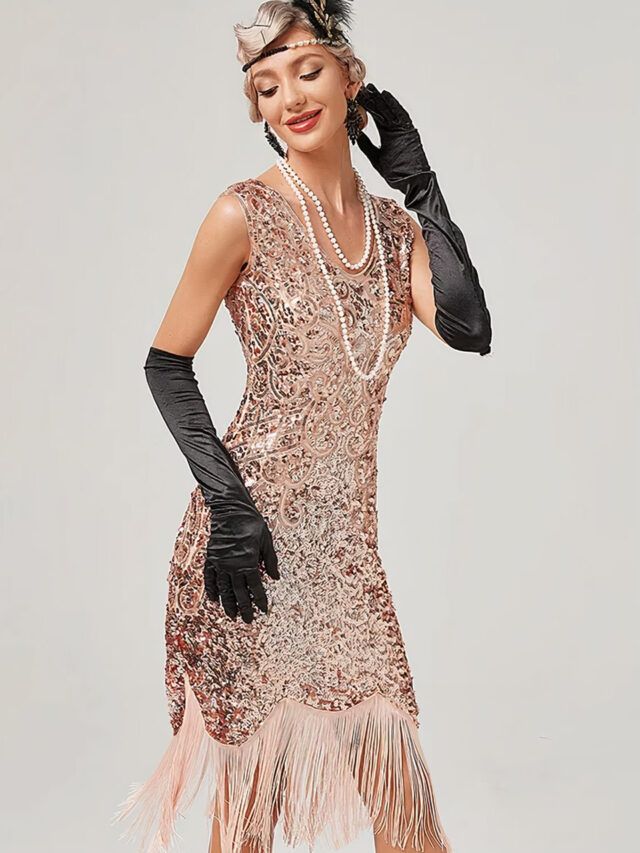 Photo d'une femme portant une robe rose entièrement pailletée style années 20 avec franges roses en bas, un collier de perles blanches, de longs gants noirs satinés, un serre-tête avec plume noire et boucles d'oreilles noires, le tout sur un fond gris