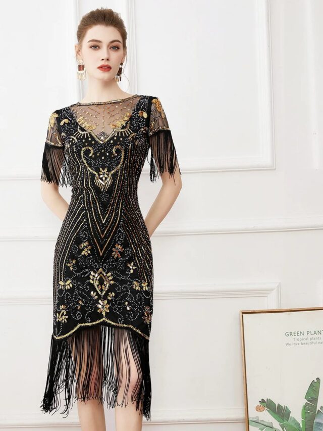 Photo d'une jeune femme debout devant un mur blanc à encadrements de même couleur, portant une robe style années 20 noire à paillettes dorées et franges sur le bas et aux manches, le haut à encolure ronde est un voile transparent comme les manches