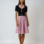Photo d'une robe plissée années 50 à col, bords manches et jupon roses à pois noirs et haut noir, portée par une jeune femme brune sur un fond gris