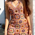 Photo d'une jeune femme debout devant un mur portant une mini robe années 70 au crochet style patchwork à dominance orange