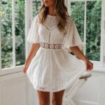 Photo d'une femme portant une robe bohème chic en coton à broderies, blanche, courte, avec des sandales, devant des fenêtres.