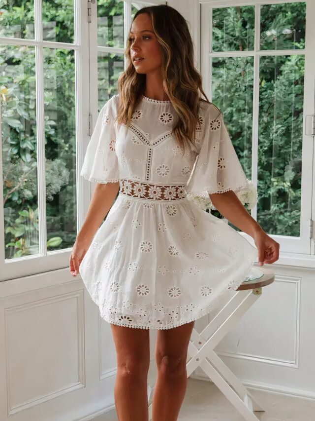 Photo d'une femme portant une robe bohème chic en coton à broderies, blanche, courte, avec des sandales, devant des fenêtres.