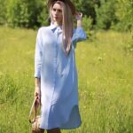 On voit une femme blonde dans un champs de hautes herbes qui porte une robe chemise en lin bleu.
