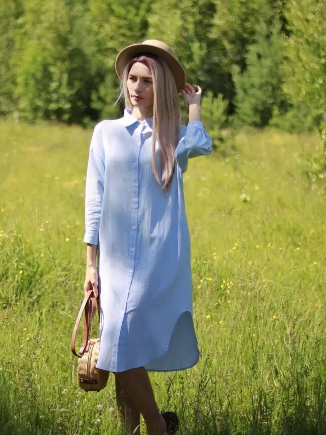 On voit une femme blonde dans un champs de hautes herbes qui porte une robe chemise en lin bleu.