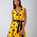 Photo d'une robe style années 50 jaune à gros pois noirs décolleté en V sans manches et ceinture noire nouée devant portée par une jeune femme brune, le tout sur un fond gris