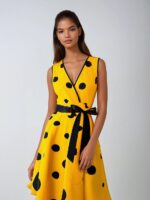 Photo d'une robe style années 50 jaune à gros pois noirs décolleté en V sans manches et ceinture noire nouée devant portée par une jeune femme brune, le tout sur un fond gris