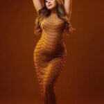 Une femme enceinte aux cheveux longs porte une robe dorée moulante et longue. Elle a les bras levés