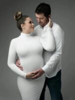 Un couple formé par un homme brun et une femme enceinte portant un chignon. La femme porte une robe blanche moulante avec un col haut. L'homme porte une chemise blanche et un pantalon noir. La femme est dans les bras de l'homme.