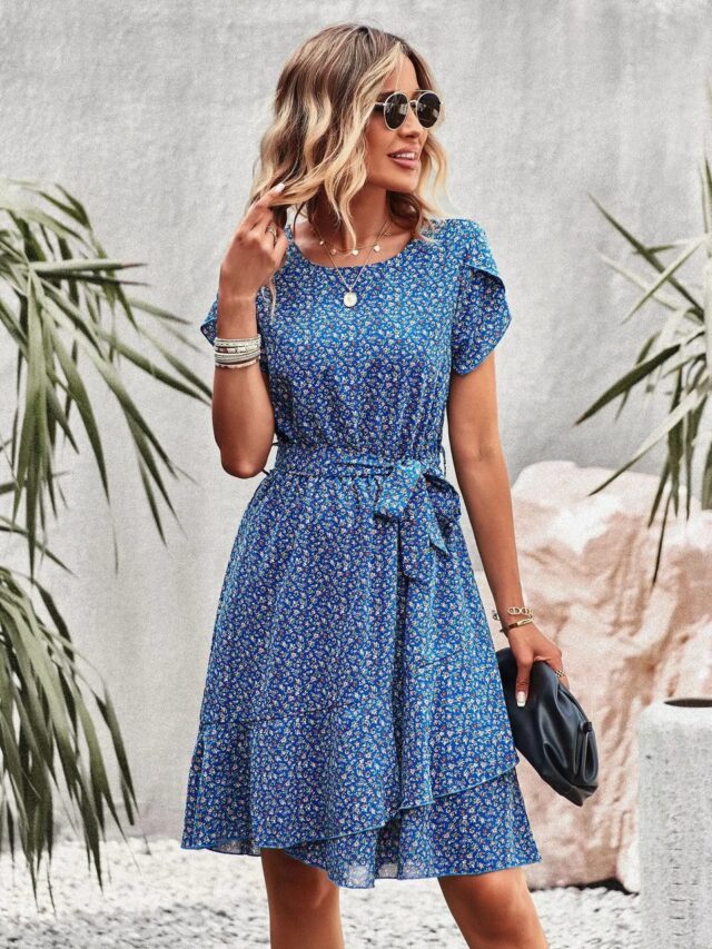 Photo d'une femme portant une robe fleurie bleue, ceinturée, à manches courtes avec un sac pochette noir à la main et des lunettes de soleil. Elle se tient debout devant un mur en béton avec des feuilles tropicales sur les côtés et a la main dans les cheveux.