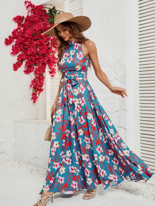 Photo d'une femme portant une robe fleurie longue élégante bleu avec un col montant, sans manches. Elle porte un chapeau de paille et semble marcher devant un mur blanc avec de grosses fleurs rouges au second plan.
