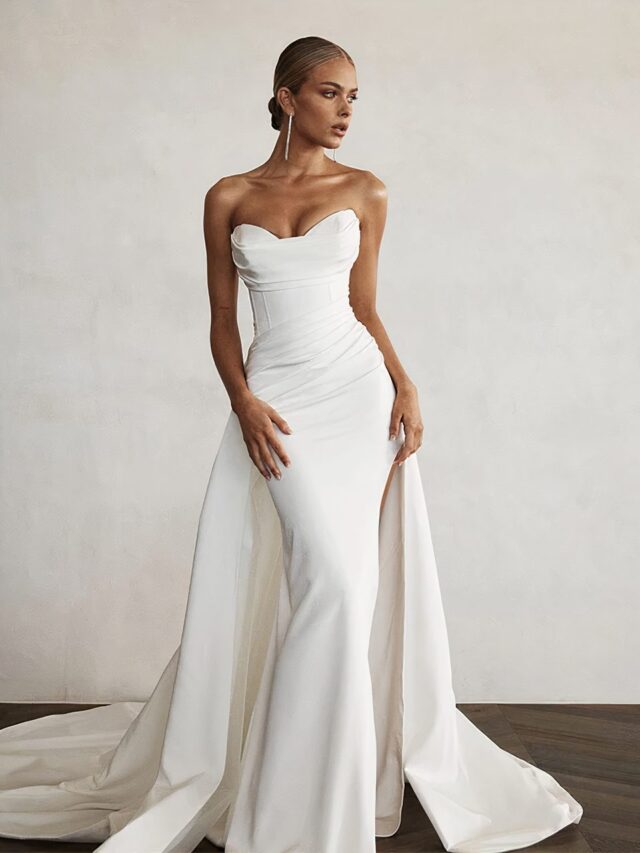 Photo d'une femme portant une robe de mariée blanche bustier corset de forme sirène avec une traîne sur un fond gris et un sol marron