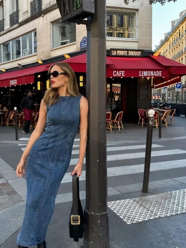 Photo d'une femme portant une robe fourreau en jean adossée à un poteau dans la rue, devant un café.